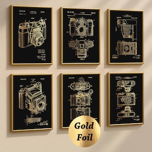 May include: Six framed vintage camera patent prints. Each print features a gold-coloured illustration of a camera on a black background, set within a gold frame. The prints are arranged in a grid pattern on a neutral-coloured wall.