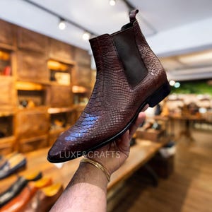 May include: A brown leather Chelsea boot with a textured, reptile-like pattern. The boot has a pointed toe and elastic side panels. The sole is black. The text "LUXE&CRAFTS" is visible on the side of the boot.