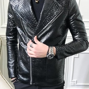 Pu&ograve; includere: Giacca nera in similpelle strutturata con motivo a pelle di serpente, chiusura con zip e colletto a revers. Indossata sopra un dolcevita nero. Sono visibili un orologio e un anello argentati.