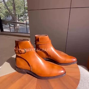 May include: Two pairs of brown leather ankle boots with buckle straps. The boots are displayed on a wooden table, with the text "NUXES & CRAFT" visible on one boot. The boots have a classic design and a polished finish.