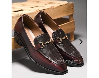 May include: A pair of burgundy leather loafers with a square toe and a crocodile-textured vamp. The shoes have a gold-colored metal buckle and a white stitched sole. The text "LUXES&CRAFTS" is visible on the right shoe.