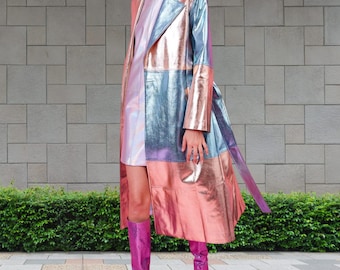 May include: A long, iridescent, multi-colored raincoat with a belt. The raincoat is made of a shiny, metallic fabric and has a patchwork design. The raincoat is worn over a white dress and paired with pink boots.