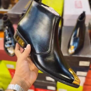 May include: Black leather ankle boot with a gold-colored metal toe cap and heel accent. The boot has a sleek, pointed toe design. The text "LUXES&CRAFTS" is visible in the image.