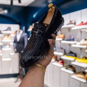 Könnte beinhalten: Schwarzer Lackleder-Loafer mit Reptilienmuster auf dem Vorderblatt und einer silberfarbenen Schnalle. Der Absatz hat silberfarbene Nieten. Der Schuh wird von einem Holzschuhspanner gehalten. Der Text "LUXE&CRAFTS" ist sichtbar.