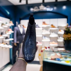 Op de afbeelding: Een zwarte leren laars met een slangenleerpatroon wordt getoond. De laars heeft een rits en een puntige neus. Het logo van de winkel, "LUXE&CRAFTS", is zichtbaar. De achtergrond toont een schoenenwinkel.