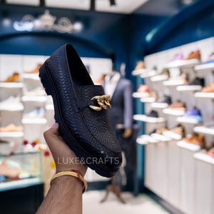 Puede incluir: Mocasín de cuero negro con textura de piel de serpiente y detalle de cadena dorada. El zapato tiene una suela gruesa y lo sostiene una persona. El nombre de la tienda, LUXE & CRAFTS, es visible en la imagen.