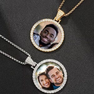 Puede incluir: Dos collares con colgantes redondos con fotos en el interior. Uno es dorado con una cadena dorada, el otro es plateado con una cadena plateada. Ambos colgantes tienen un borde de pequeñas piedras transparentes. Las fotos son de personas sonriendo.