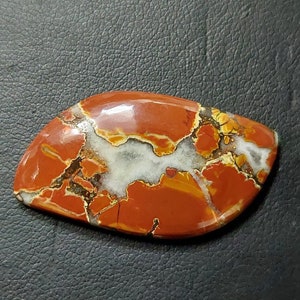 Puede incluir: Un cabujón de ágata rojo, blanco y marrón con una superficie pulida. La piedra tiene un patrón único de colores y vetas en espiral.