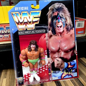 May include: An action figure of the Ultimate Warrior, packaged on a card with the WWF logo and a photo of the wrestler. The figure is in a clear plastic case. The card has red, white, and blue colors. The figure is wearing green trunks and white boots.