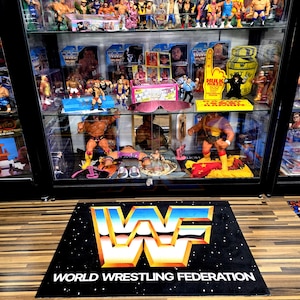 May include: A black rug with the World Wrestling Federation logo in blue, orange, and white, and the text "WORLD WRESTLING FEDERATION". A glass display case filled with wrestling action figures and memorabilia is behind the rug.