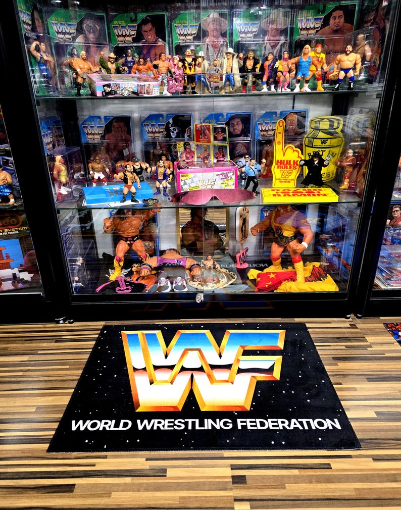 May include: A black rug with the World Wrestling Federation logo in blue, orange, and white, and the text "WORLD WRESTLING FEDERATION". A glass display case filled with wrestling action figures and memorabilia is behind the rug.