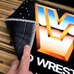 May include: Black rectangular mat with a white stitched border. The mat features a large, stylized gold and blue letter "W" and the words "WORLD WRESTLING". The mat is partially rolled up, revealing the underside.