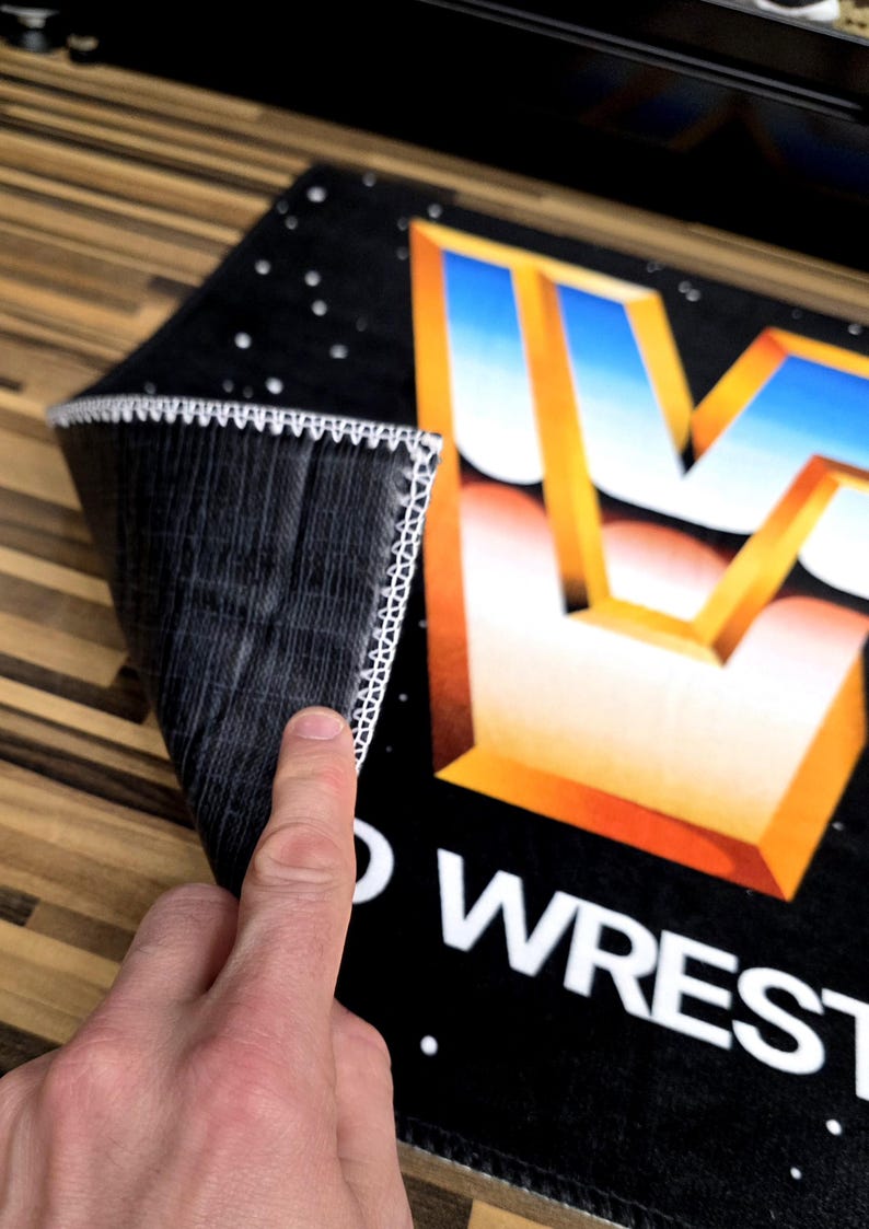May include: Black rectangular mat with a white stitched border. The mat features a large, stylized gold and blue letter "W" and the words "WORLD WRESTLING". The mat is partially rolled up, revealing the underside.