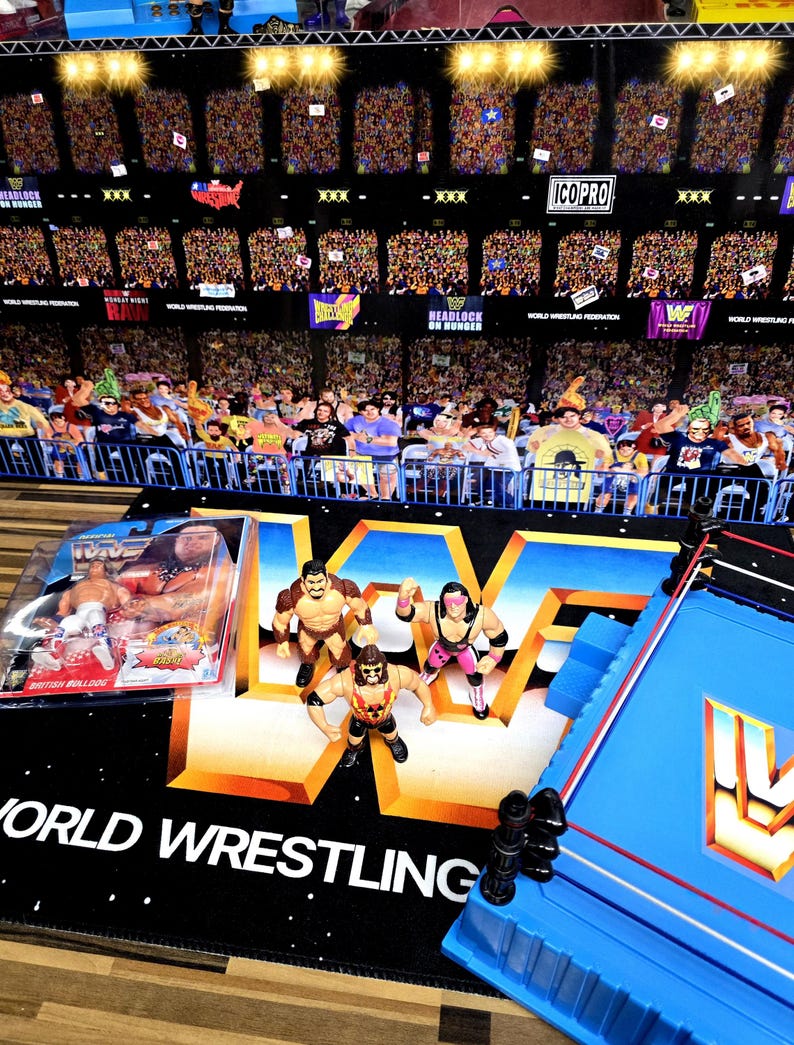 May include: A collection of vintage wrestling action figures and a miniature wrestling ring. The figures are posed on a black mat with the words "World Wrestling" in white. A packaged figure is also visible, along with a blue wrestling ring.