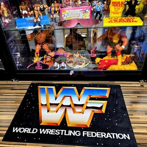 May include: Black rectangular rug with the World Wrestling Federation logo in white, blue, and orange. The text "WORLD WRESTLING FEDERATION" is below the logo. The background has a starry pattern. A glass display case with wrestling figures is behind the rug.