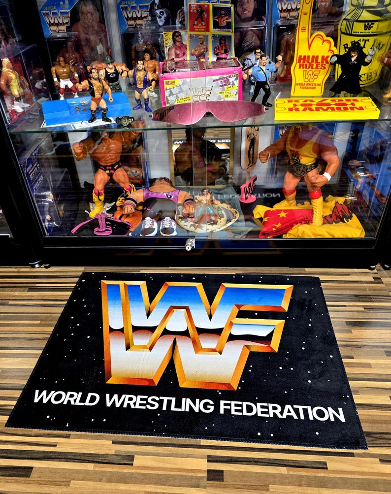 May include: Black rectangular rug with the World Wrestling Federation logo in white, blue, and orange. The text "WORLD WRESTLING FEDERATION" is below the logo. The background has a starry pattern. A glass display case with wrestling figures is behind the rug.