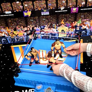 May include: A blue wrestling ring toy set with four action figures. The ring has red and white ropes, and the background shows a crowd in a stadium. The set includes a small table with the text "World Wrestling Federation".
