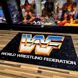 May include: Black rectangular rug with the WWF logo in white, gold, and blue. The words "WORLD WRESTLING FEDERATION" are printed in white below the logo. The rug is on a wooden floor, and a display case with wrestling figures is in the background.