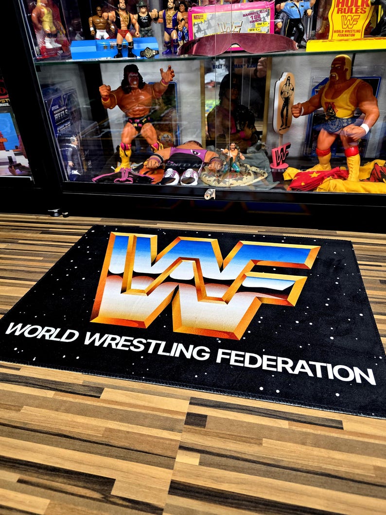 May include: Black rectangular rug with the WWF logo in white, gold, and blue. The words "WORLD WRESTLING FEDERATION" are printed in white below the logo. The rug is on a wooden floor, and a display case with wrestling figures is in the background.