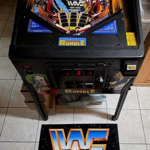 May include: A black and blue WWF Royal Rumble pinball machine with a colorful playfield featuring wrestling characters. The machine has a black cabinet and legs, with the WWF logo on a mat in front. The text "DATA EAST PRESENTS THE ROYAL RUMBLE" is visible.