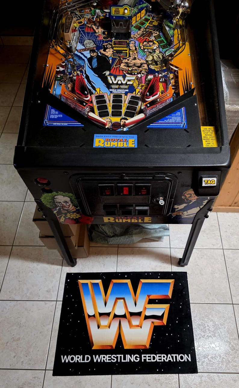 May include: A black and blue WWF Royal Rumble pinball machine with a colorful playfield featuring wrestling characters. The machine has a black cabinet and legs, with the WWF logo on a mat in front. The text "DATA EAST PRESENTS THE ROYAL RUMBLE" is visible.
