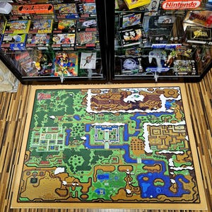 Könnte beinhalten: Ein rechteckiger Teppich mit einer detaillierten Karte einer Videospielwelt, mit grünen, braunen und blauen Bereichen. Der Teppich hat einen hellbraunen Rahmen und liegt auf einem Holzboden. Im Hintergrund befindet sich eine Glasvitrine mit Videospielen.