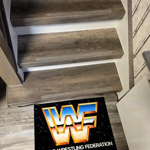 May include: Wooden staircase with three steps, featuring a gray-brown wood grain pattern. A black mat at the base displays the World Wrestling Federation logo in blue, orange, and white, with the text "WORLD WRESTLING FEDERATION".