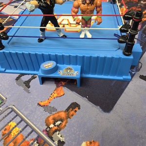May include: A blue plastic wrestling ring toy with two action figures. One figure is dressed in black, the other in green and pink. The ring has red, white, and blue ropes. A small blue table with the text "Wrestlemania" is in the foreground.