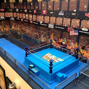 May include: A blue wrestling ring with red ropes and a blue mat. The ring is surrounded by a black metal barricade. The ring is set up in a miniature wrestling arena with a backdrop of a crowd of people. The backdrop is a printed image of a wrestling arena with a crowd of people.