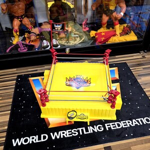 May include: A yellow World Wrestling Federation (WWF) wrestling ring toy with red corner posts and white, red, and blue ropes. The ring sits on a black mat with the words "WORLD WRESTLING FEDERATION" in white.