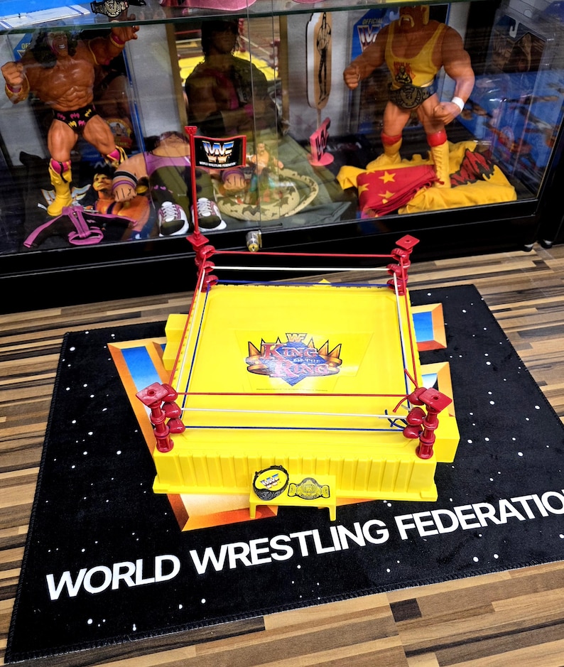 May include: A yellow World Wrestling Federation (WWF) wrestling ring toy with red corner posts and white, red, and blue ropes. The ring sits on a black mat with the words "WORLD WRESTLING FEDERATION" in white.