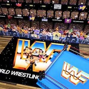 May include: A toy wrestling ring with a blue base and red and white ropes. A black mat with the WWF logo and wrestling figures are in the foreground. The background shows a crowd of people in an arena.