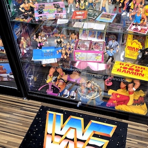 May include: A display case filled with vintage WWF (World Wrestling Federation) action figures and memorabilia. The case features various wrestlers, action figure packaging, and a black mat with the WWF logo and text.