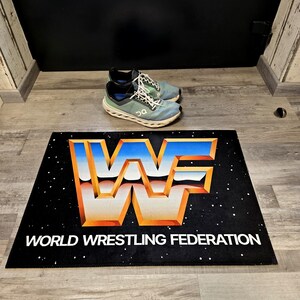May include: Black rectangular doormat with the World Wrestling Federation logo in orange, blue, and white. The words "WORLD WRESTLING FEDERATION" are printed in white below the logo. A pair of blue and green athletic shoes are on the floor.