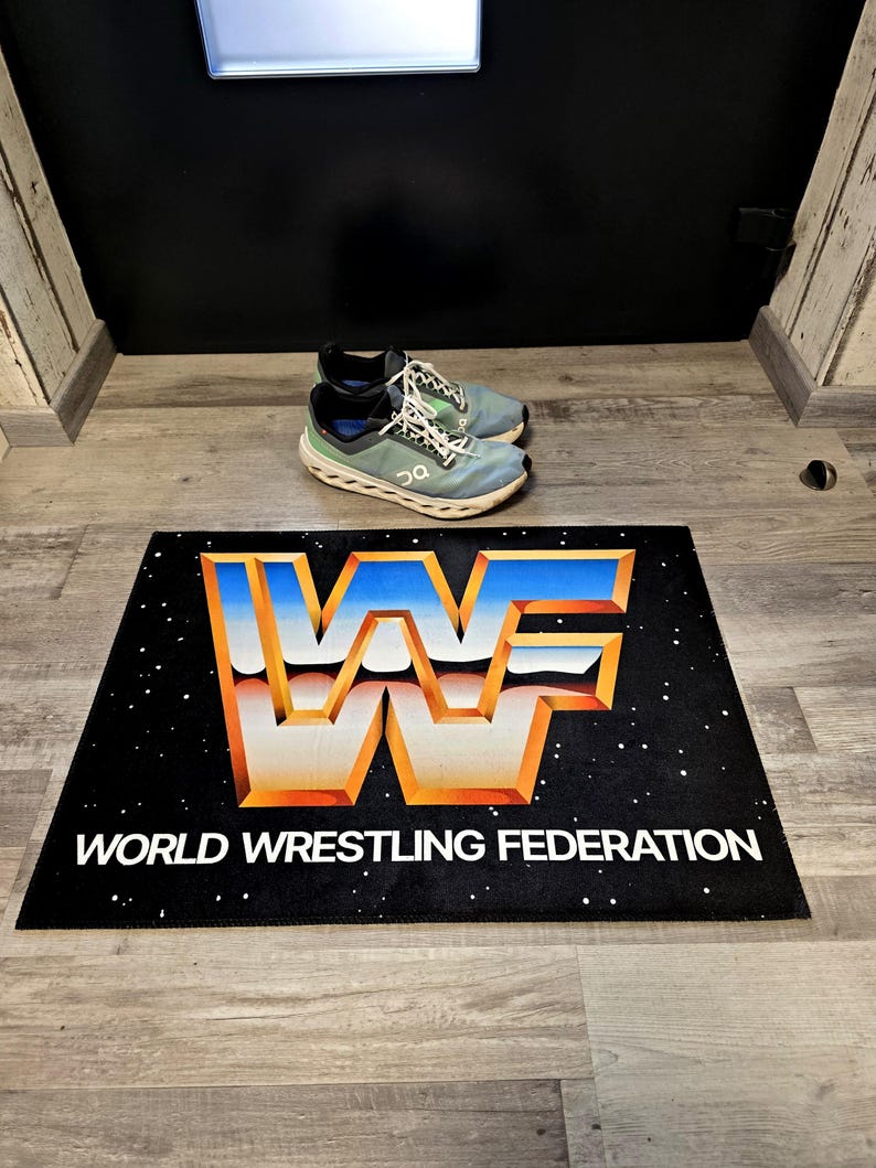 May include: Black rectangular doormat with the World Wrestling Federation logo in orange, blue, and white. The words "WORLD WRESTLING FEDERATION" are printed in white below the logo. A pair of blue and green athletic shoes are on the floor.