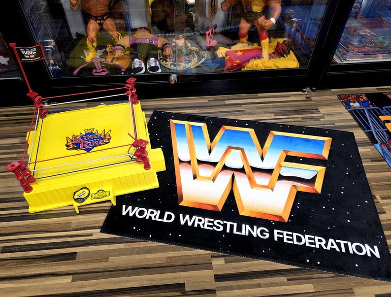 May include: A yellow wrestling ring toy with red corner posts and ropes. A black mat with the WWF logo and text "WORLD WRESTLING FEDERATION" is next to the ring. The ring has the text "King of the Ring".