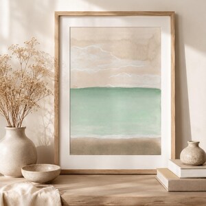Pode incluir: Uma impressão de arte de parede vertical emoldurada apresentando uma paisagem abstrata em aquarela de uma praia com um primeiro plano de areia marrom claro, um oceano verde menta e um céu bege com finas linhas brancas horizontais.