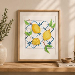 Pode incluir: Uma impressão de arte emoldurada com uma ilustração em aguarela de três limões amarelos com folhas verdes dispostos sobre um padrão de azulejos geométricos azuis e brancos. A obra está numa moldura de madeira clara com passe-partout.