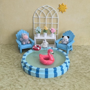 May include: A miniature pool scene with a pink flamingo float, a red life preserver, and a small penguin. Two blue chairs flank the pool, each with a stuffed animal. A white trellis and small flower pots complete the scene.