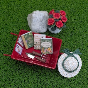 May include: A miniature gardening scene with a red wheelbarrow filled with seed packets, books, and tools. A small white stone, a bouquet of red roses, and a straw hat with a green ribbon complete the scene, all set on a green surface.