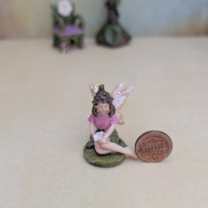 May include: A miniature fairy figurine with brown hair, a dark brown acorn-style hat, a pink shirt, and glittery wings, sitting on a green base. The figurine is positioned next to a copper-colored one-cent coin for scale, showing its small size.