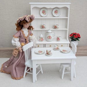 May include: A miniature dollhouse scene featuring a porcelain doll in a mauve dress, a white wooden hutch, a dining table with two chairs, and a tea set decorated with pink roses. The hutch displays plates, cups, and a teapot with floral motifs.
