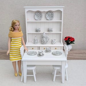 May include: A miniature white dining set featuring a hutch, table, and two chairs. A doll with blonde hair wears a yellow and white striped dress. The set includes a tea service with black and white floral patterns on plates, cups, and a teapot.