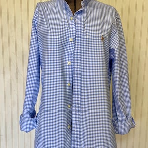 May include: A blue and white gingham button-down shirt with long sleeves and a classic collar. The shirt features a small embroidered logo on the chest and is displayed on a mannequin. The cuffs are rolled up.