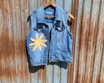 May include: A blue denim vest with a brown flower design on the front. The vest has a zipper closure and two pockets. The brand name "Cat & Jack" is visible on the label.