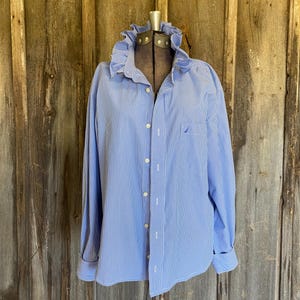 May include: A long-sleeved button-down shirt featuring a blue and white vertical pinstripe pattern. The shirt includes a prominent ruffled collar, a front button placket, and a single chest pocket with a small embroidered logo detail.