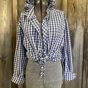 May include: A blue and white checkered button-down shirt, tied at the waist, displayed on a gray dress form. The shirt has long sleeves with rolled-up cuffs and a ruffled collar. The background is a weathered wooden wall.