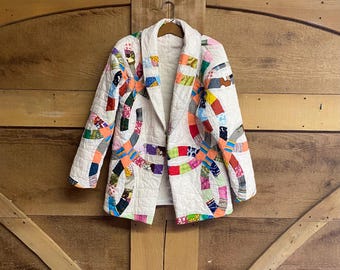 May include: A quilted jacket featuring a white base with a colorful double wedding ring pattern. The garment has long sleeves, a wide lapel collar, and a button closure. The fabric is composed of various patterned textile scraps.
