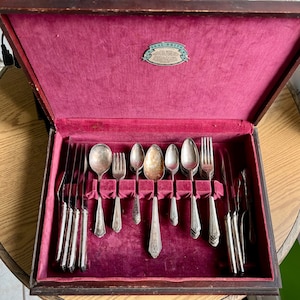 May include: An open wooden box with a red velvet interior, containing a set of vintage silver cutlery. The cutlery includes knives, forks, and spoons, all with ornate handles. The box has a label that reads "Seal-Brite" on the inside of the lid.