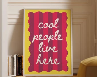 May include: A framed art print with the words "cool people live here" in white script on a red and pink striped background. The print has a yellow border and is displayed in a room with a wooden floor.